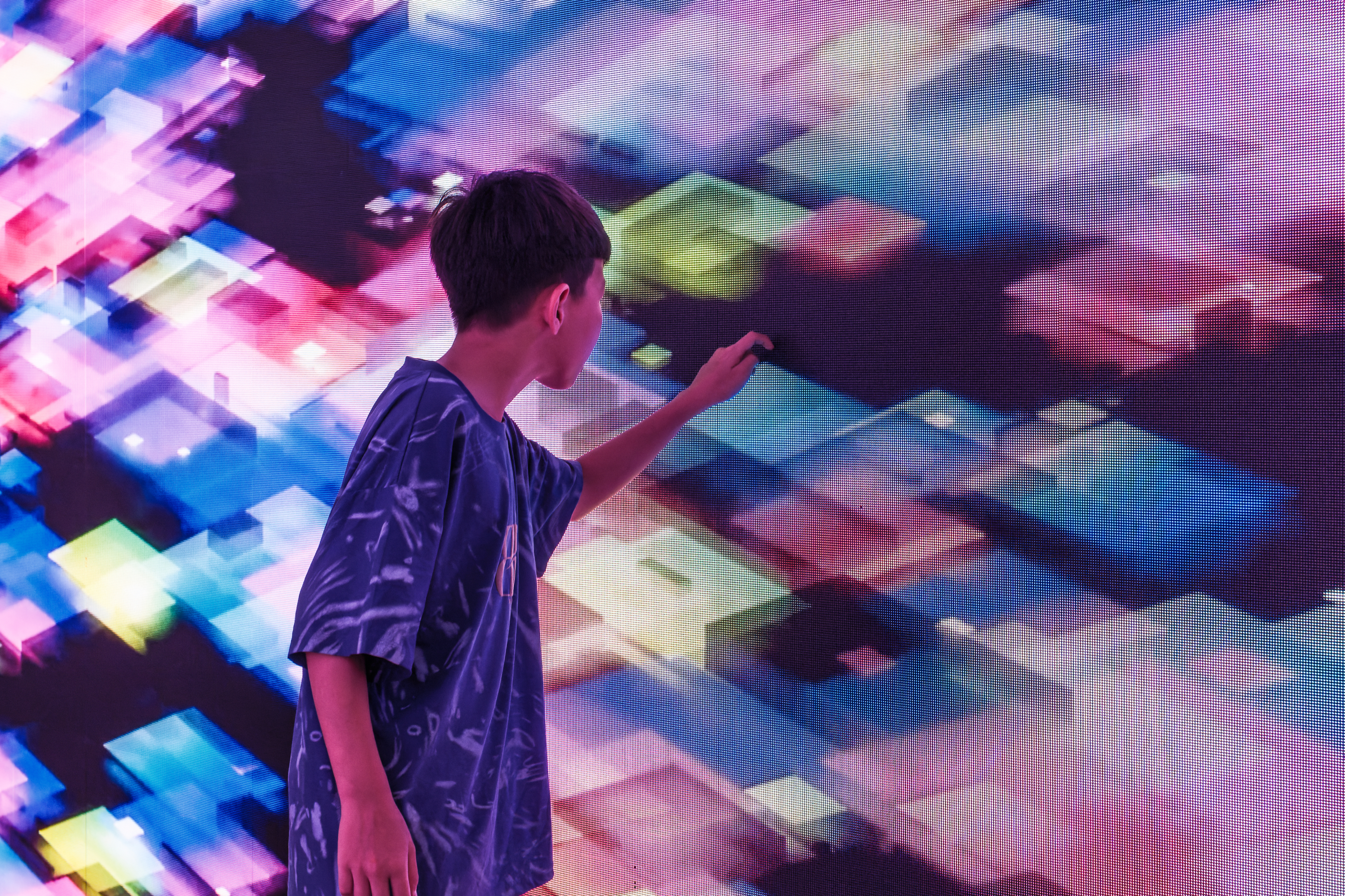 A young boy interacts with the exhibition, reaching out to touch a massive digital screen displaying bright, geometric generative art in vibrant pinks, blues, and greens.