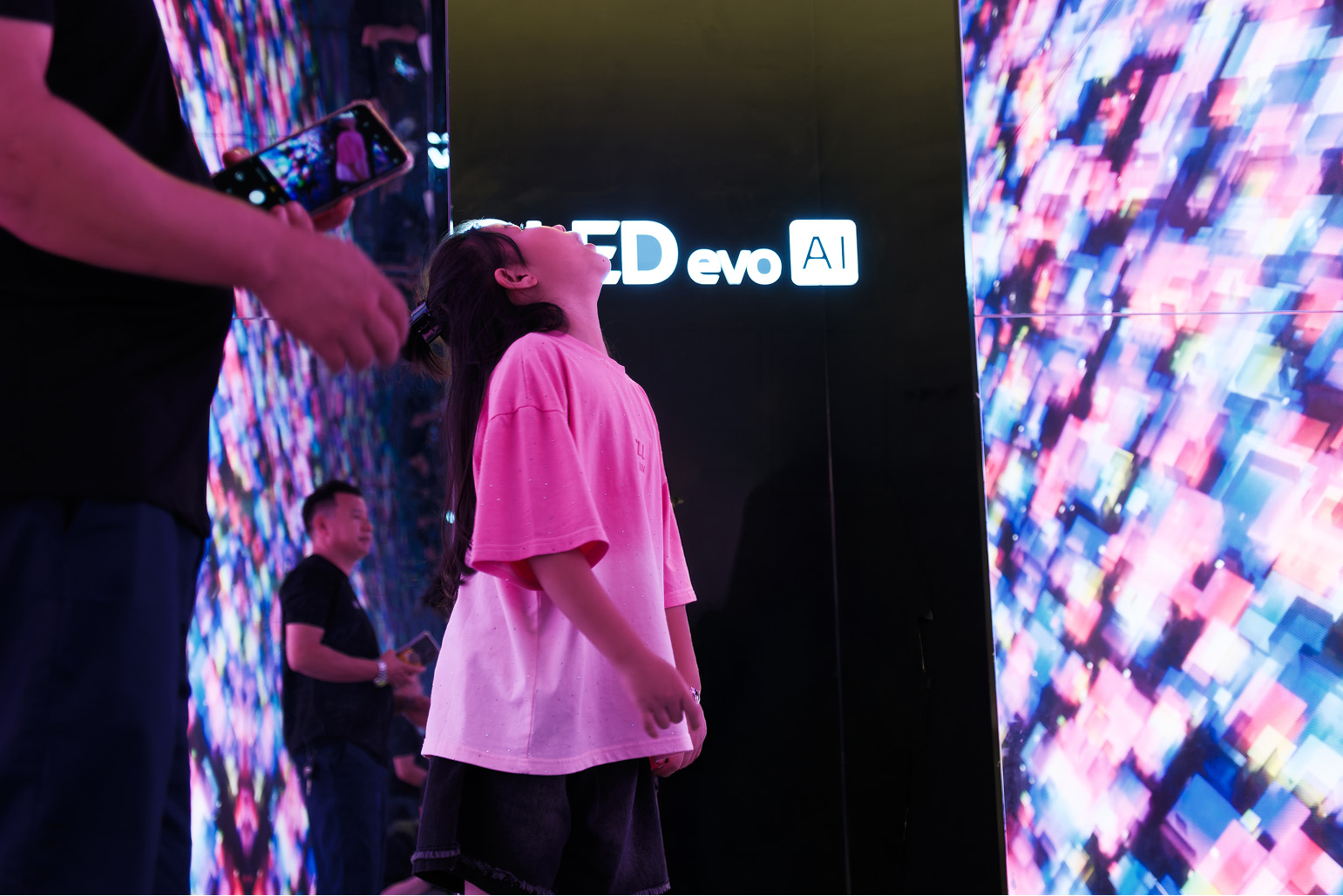 A child gazes upward at a large-scale installation showing colorful, geometric generative art. The vivid pink, blue, and purple abstract visuals illuminate her face, capturing the engaging atmosphere of the exhibition space.