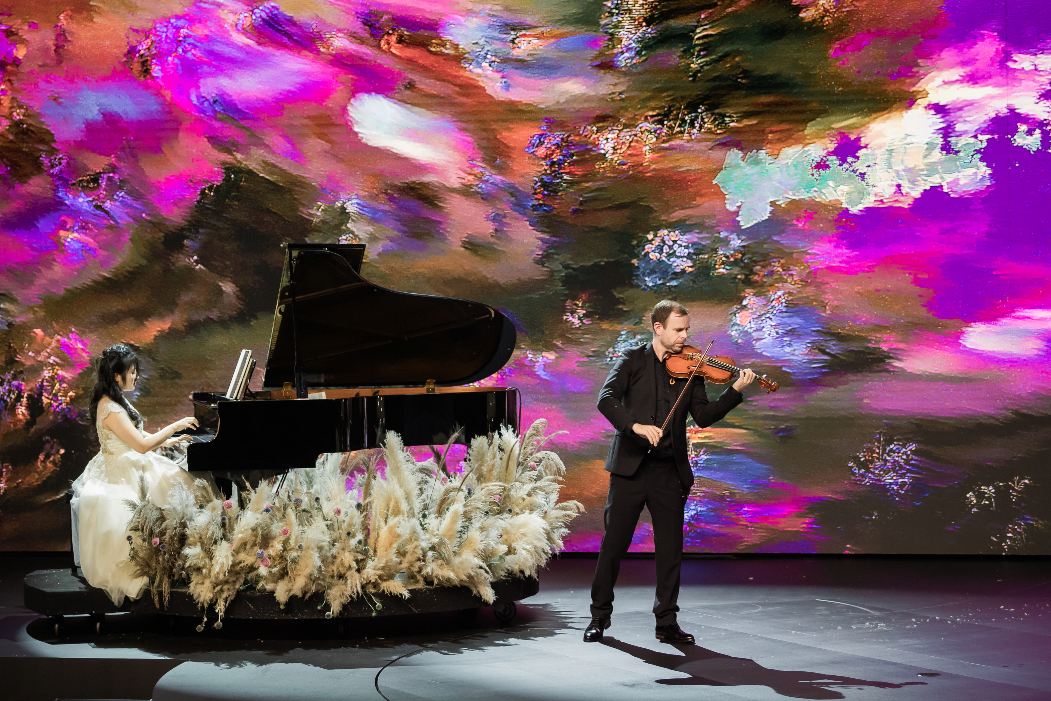 The pianist Ngô Phương Vi and the violinist Emil Kleve performing a duet. The background screen displays a textured digital canvas of vibrant pinks, purples, and greens.