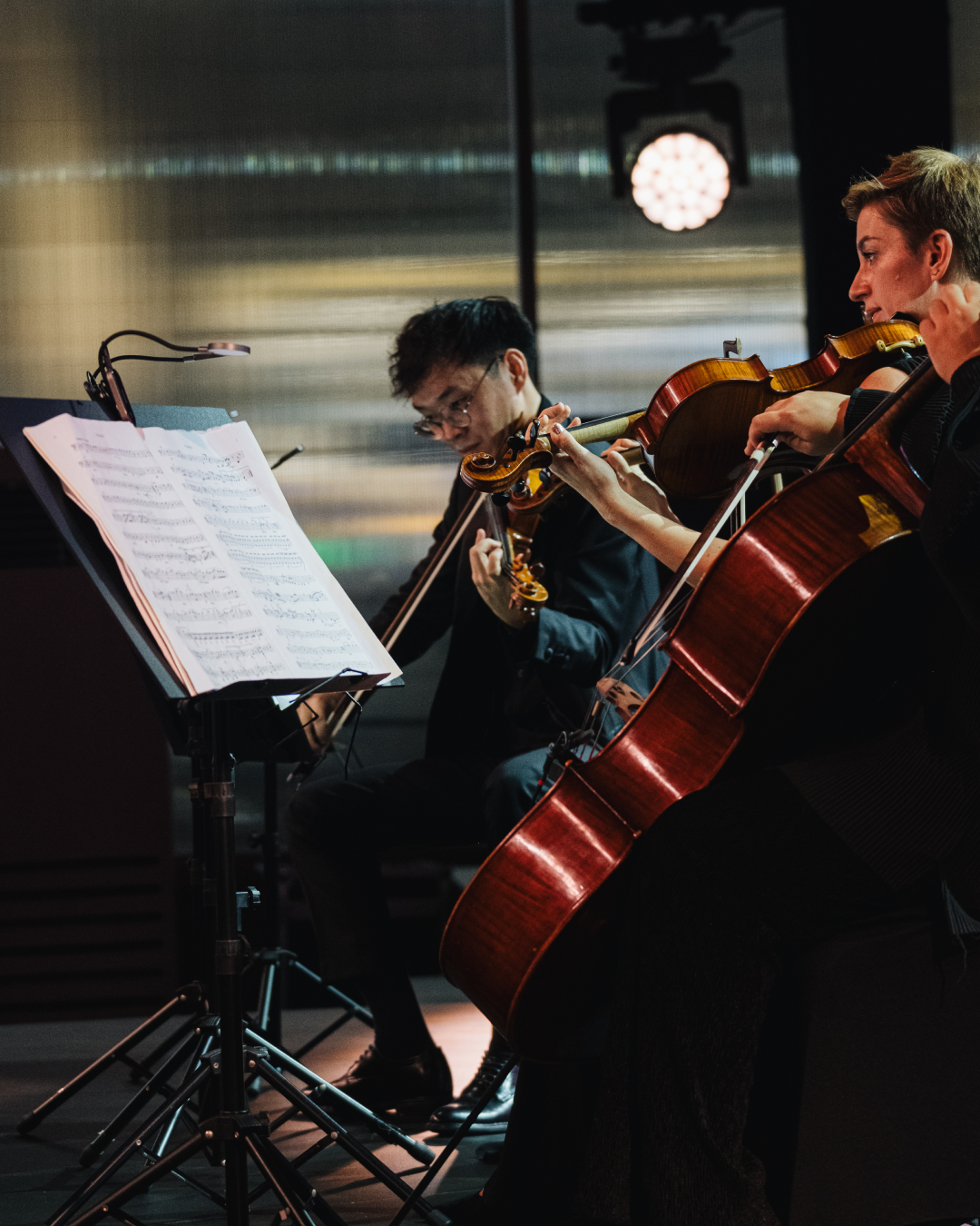 A close-up shot of the string quartet, focusing on the violinist Nguyễn Thiện Minh playing illuminated by a stand light, with a female violinist and a cello prominently in the foreground.