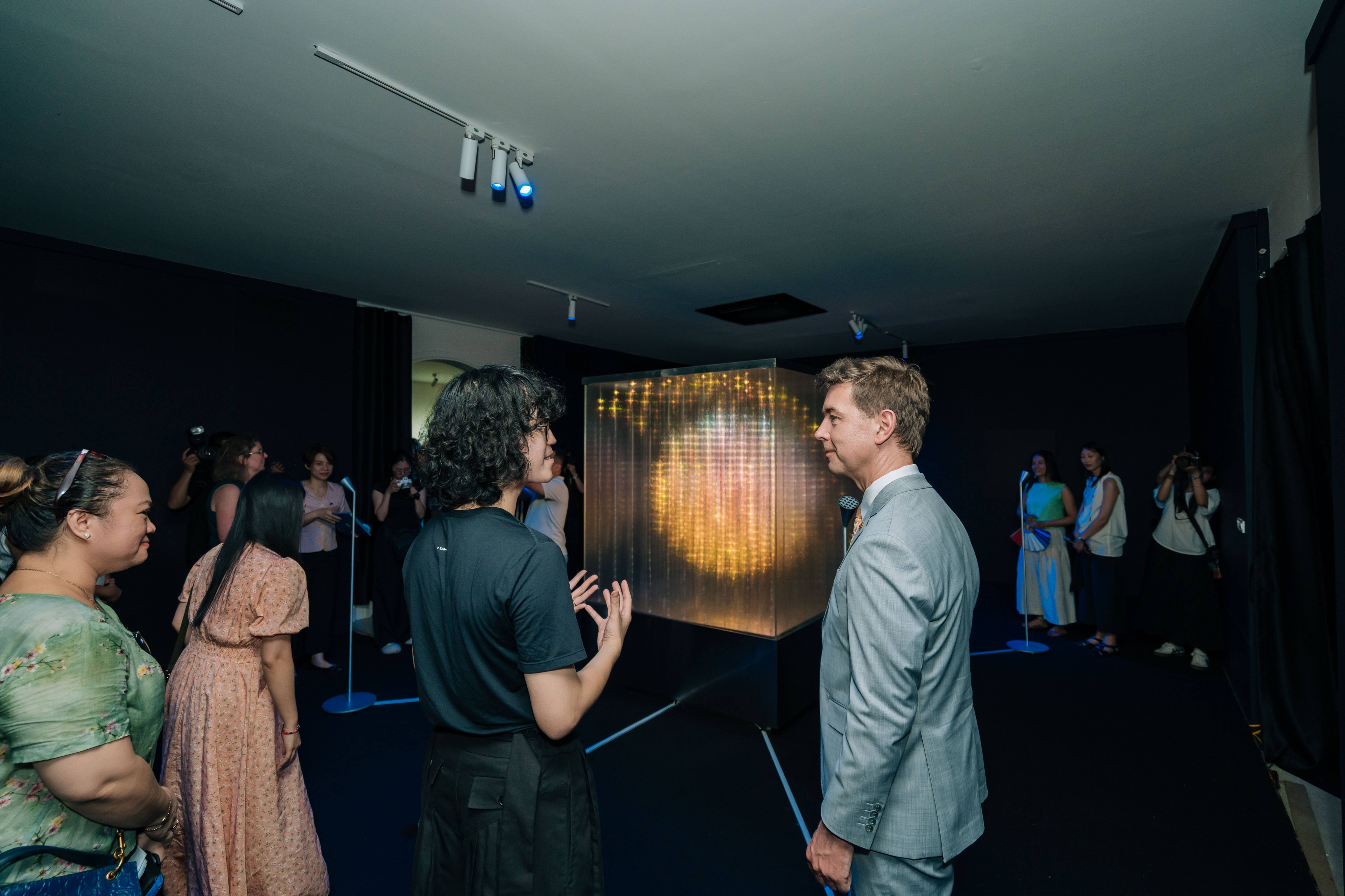 An exhibition space where Fustic. is speaking to the EU Ambassador to Vietnam in a grey suit, while a crowd observes and interacts with the installation.