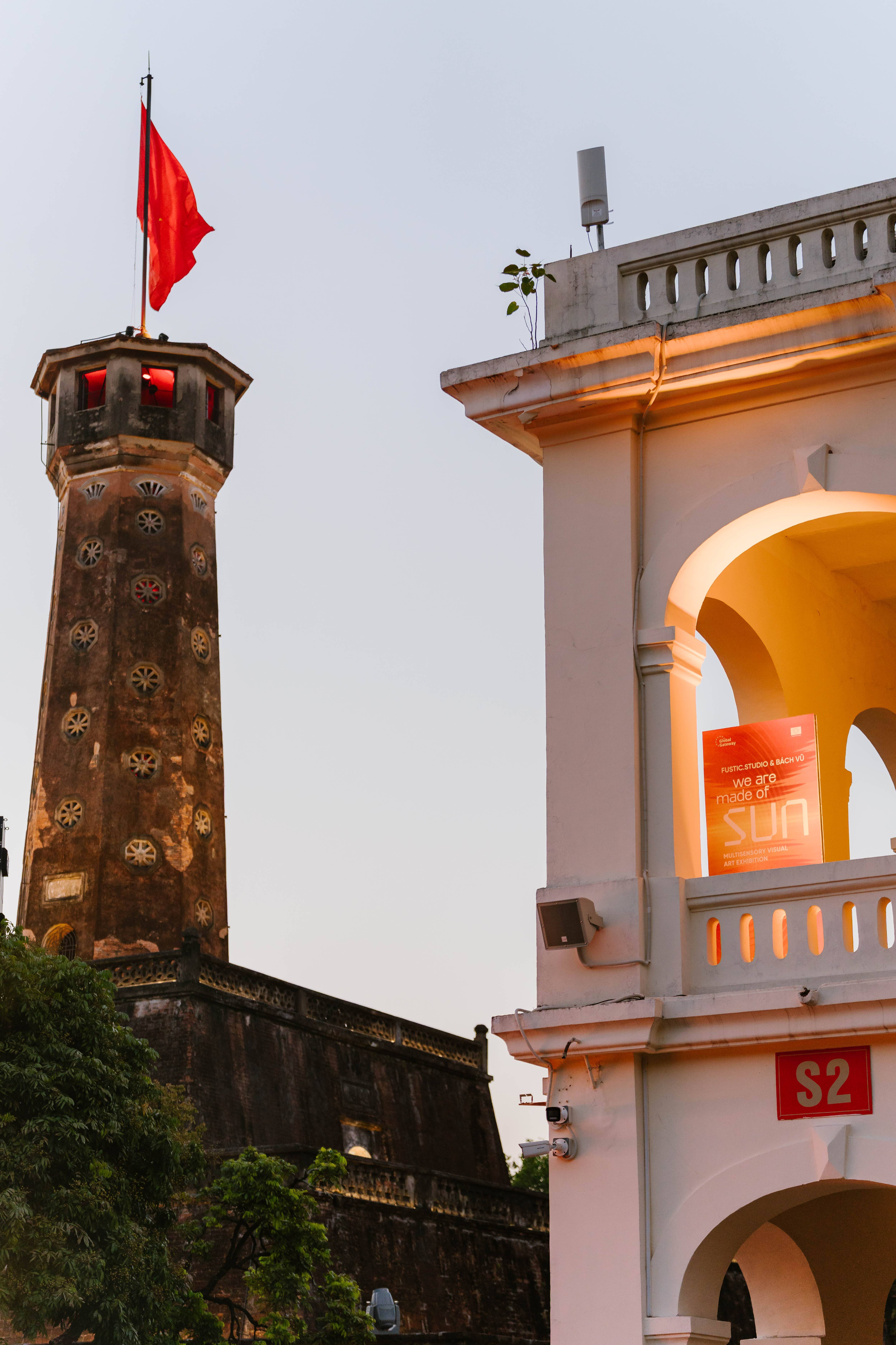 A view of the 'We Are Made of Sun' exhibition poster on a white balconied building, with a historic Hanoi Flag Tower flying the flag of Vietnam in the background at dusk.