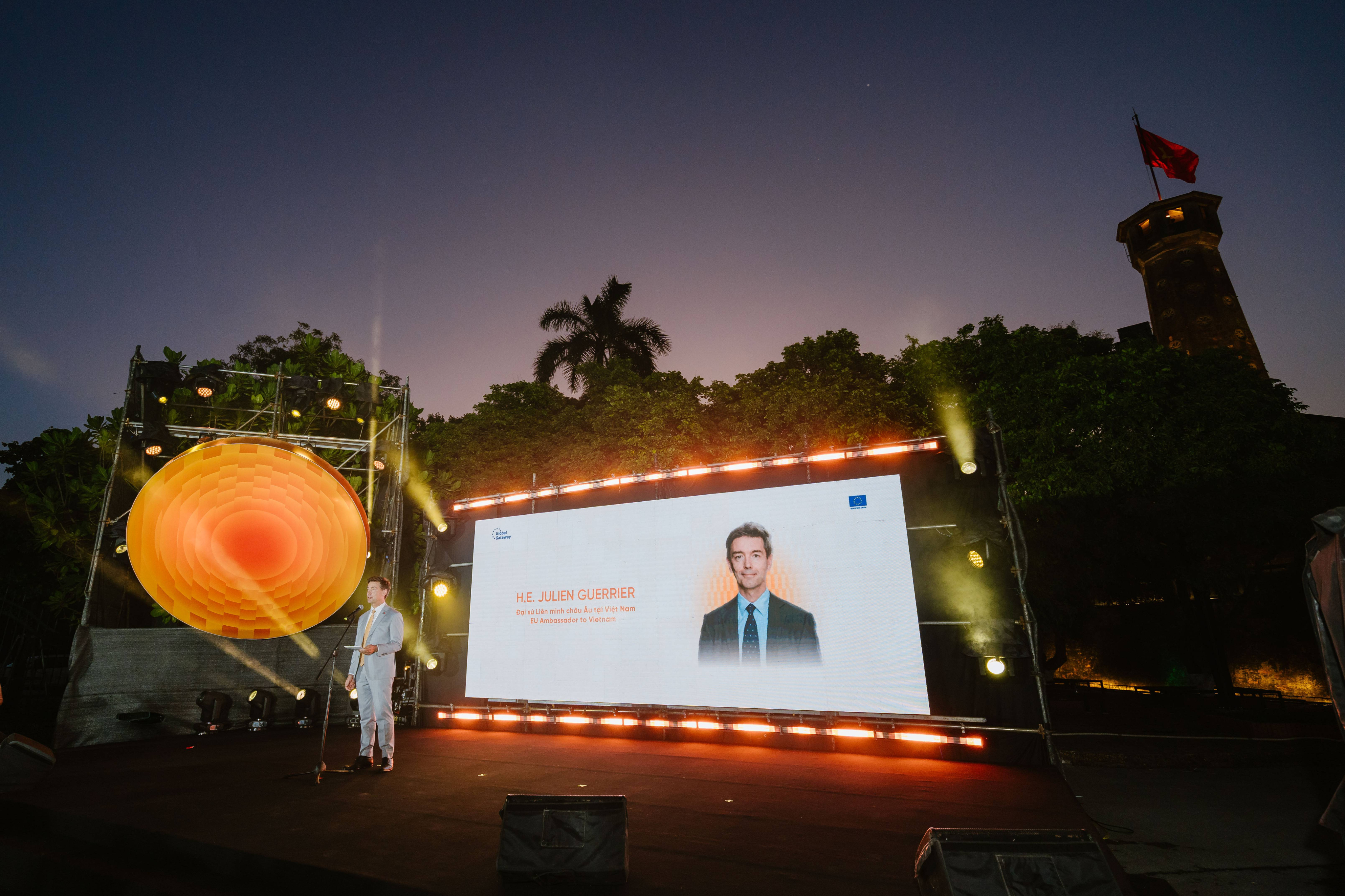The EU Ambassador to Vietnam in a light grey suit speaking at a microphone on an outdoor stage at night, alongside a large digital screen displaying his portrait and the name H.E. Julien Guerrier.